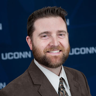 Image of Online Environmental Master's Degree Faculty Member and Course Instructor Dr. Chadwick Rittenhouse smiling in a brown suit in front of a UConn backdrop. 